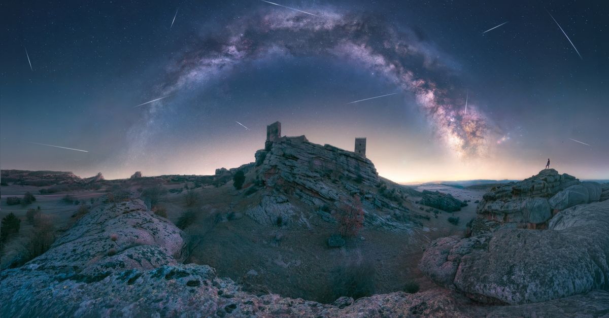 Castillo de piedra sobre una colina rocosa bajo un cielo nocturno estrellado con un brillante arco de la Vía Láctea.