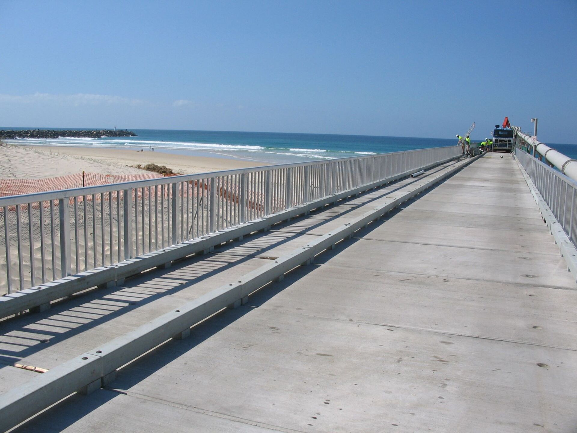 A concrete pier extends into the ocean, with metal railings and tracks on either side. Beach and ocean visible.