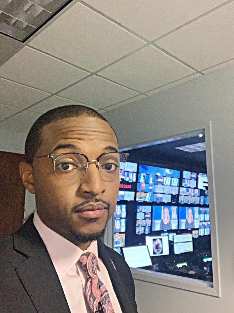 Man in glasses, suit, and tie takes a selfie in front of a window showing multiple monitors.