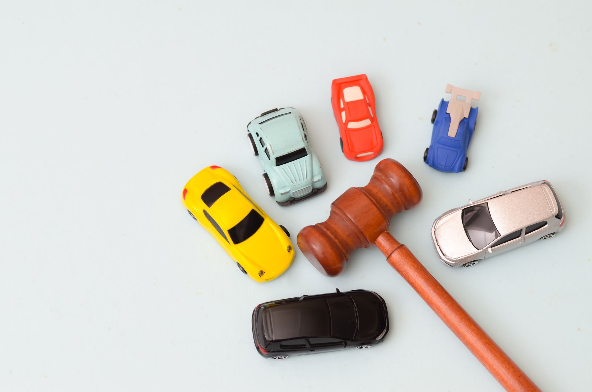 a wooden judge's gavel surrounded by six toy cars of various colors and styles on a light background.