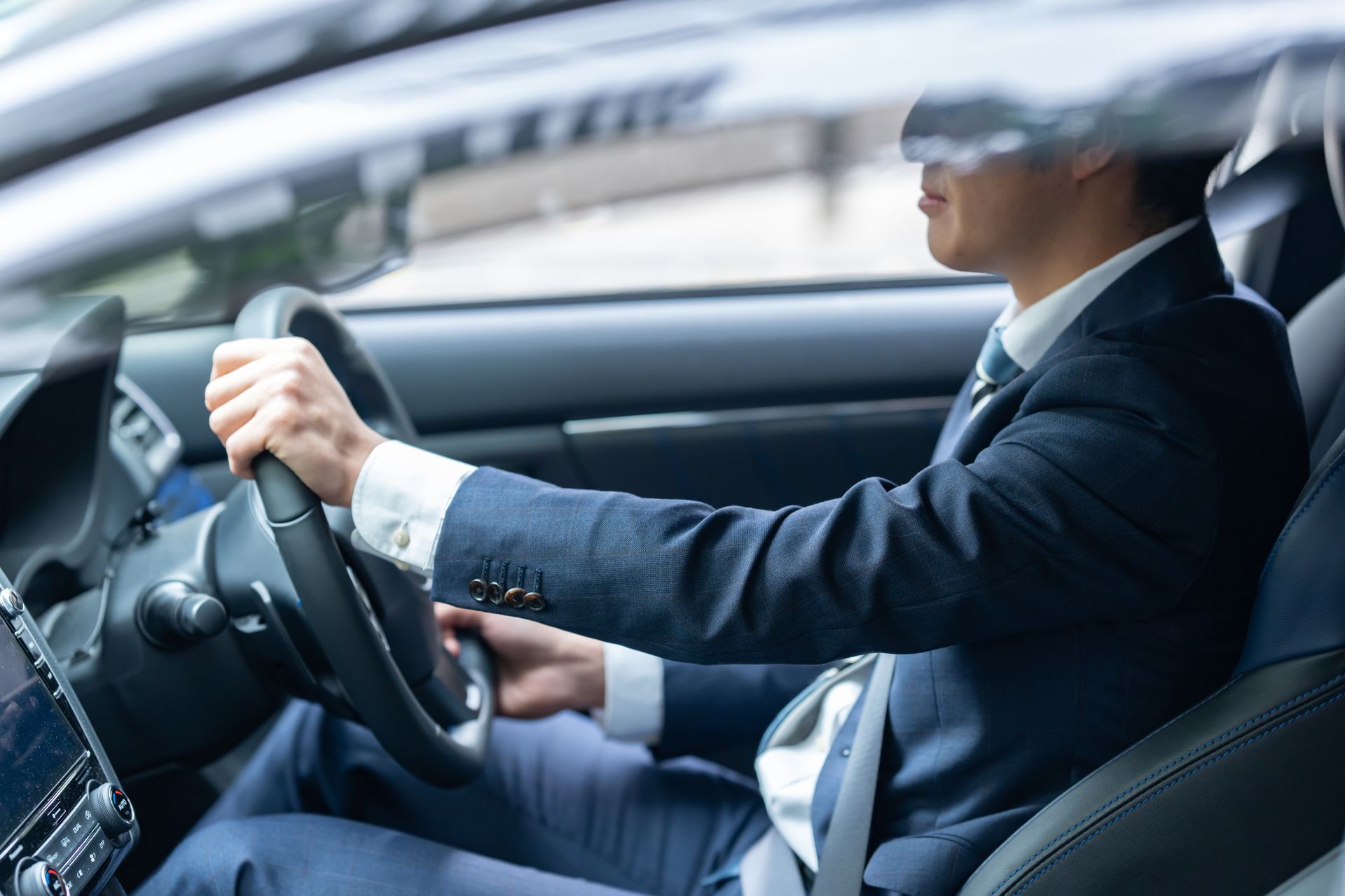 a man in a navy blue suit driving a car, focusing on his hands gripping the steering wheel and his p