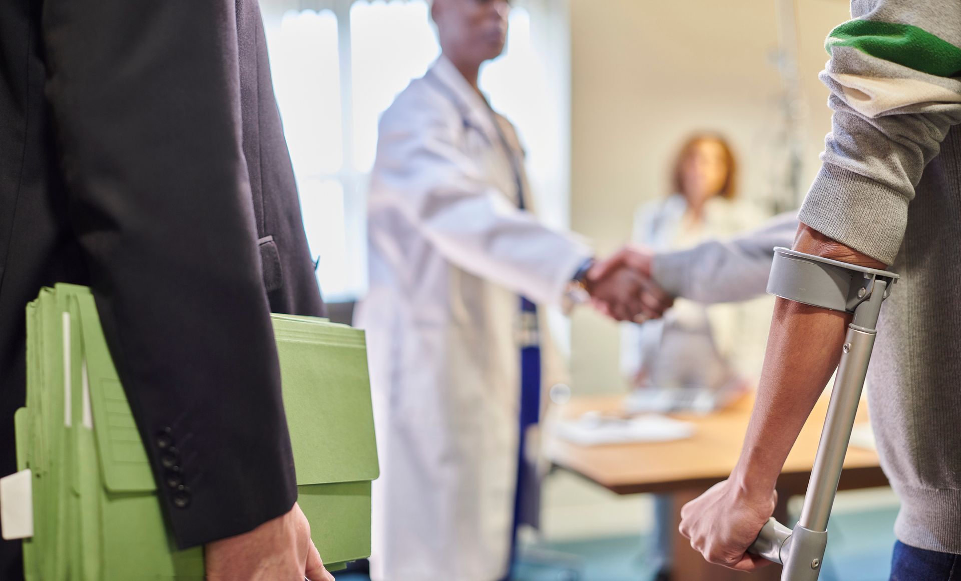 Doctor shaking hands with patient using crutch, person holding green folders nearby.