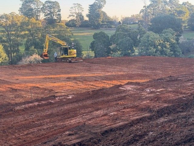 Excavator grading a large red dirt field with trees and rolling hills in the background