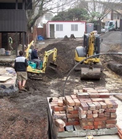 Construction workers and excavators digging a muddy residential yard beside stacked bricks.