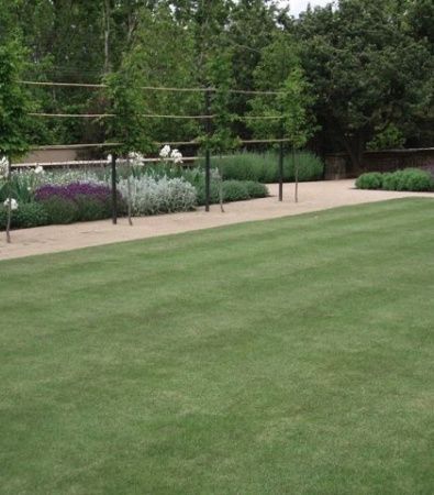 Lush garden with green lawn, flowerbeds, and a fenced walkway lined with trees and shrubs