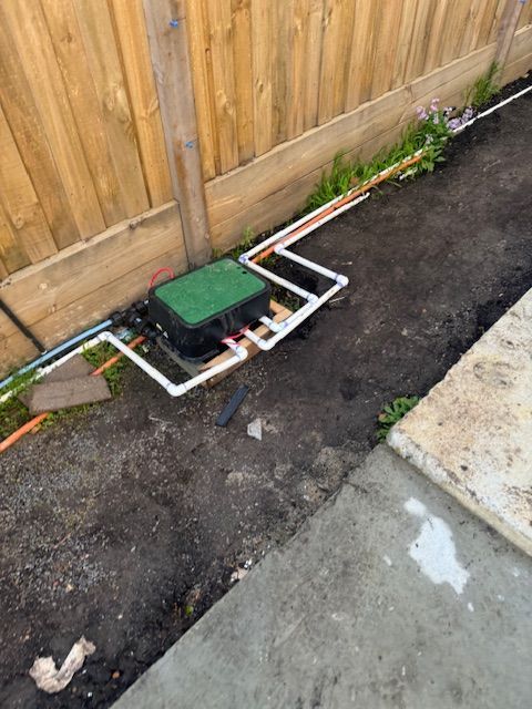 Irrigation system components: green box, white pipes, beside a wooden fence and concrete.