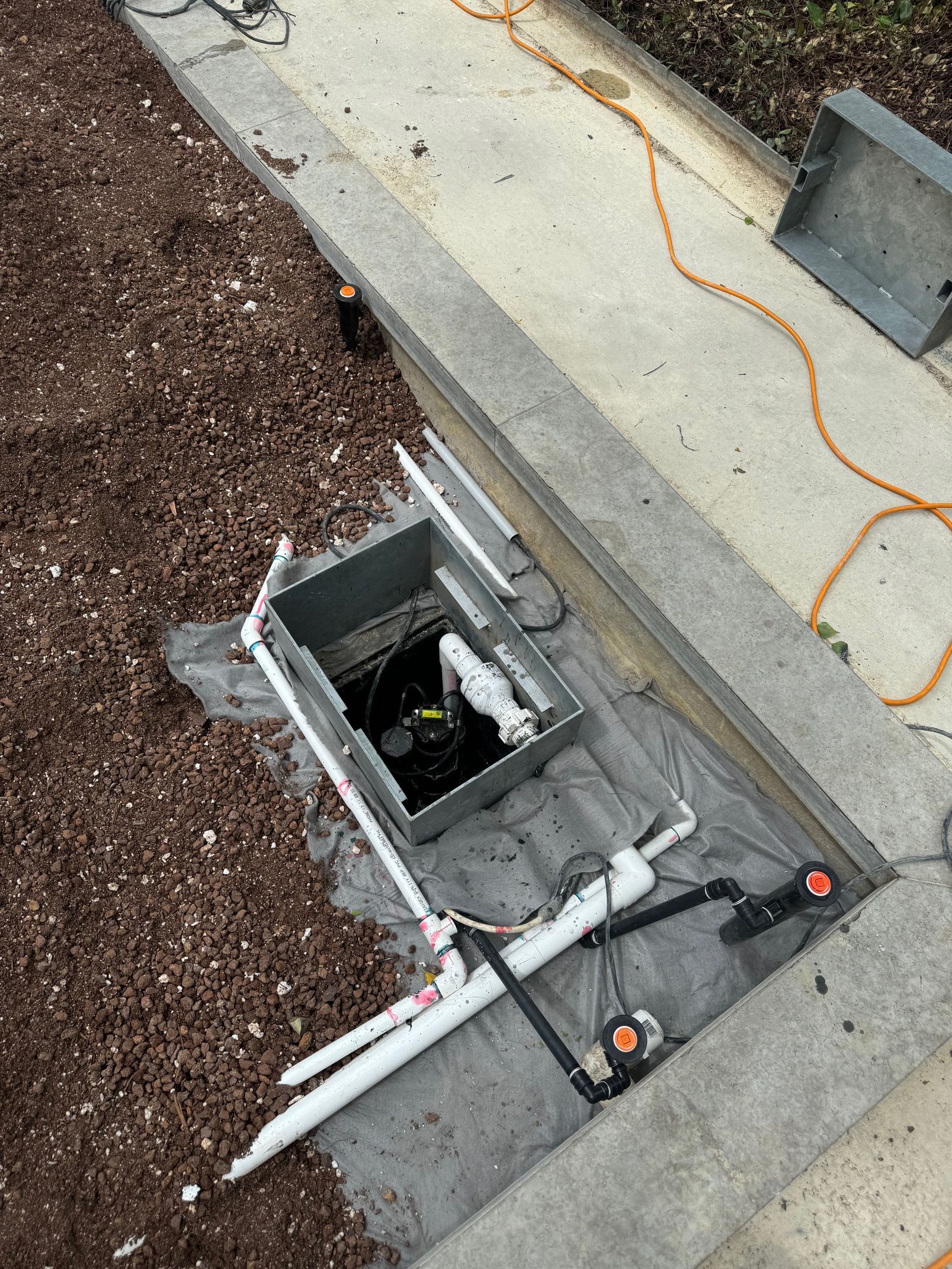 Open electrical box with exposed wires and pipes in a concrete border, covered in wood mulch.