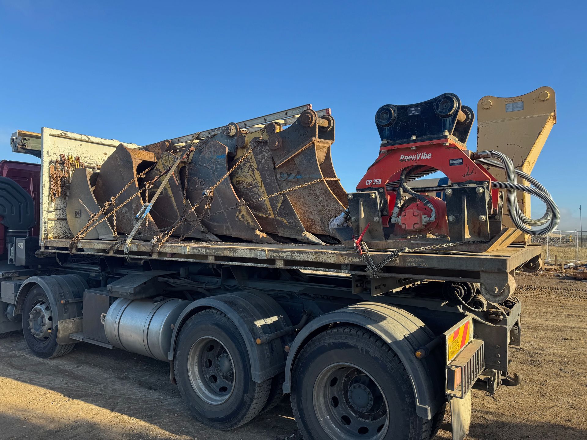 Semi-truck flatbed with large excavator attachments and a hydraulic compactor unit.