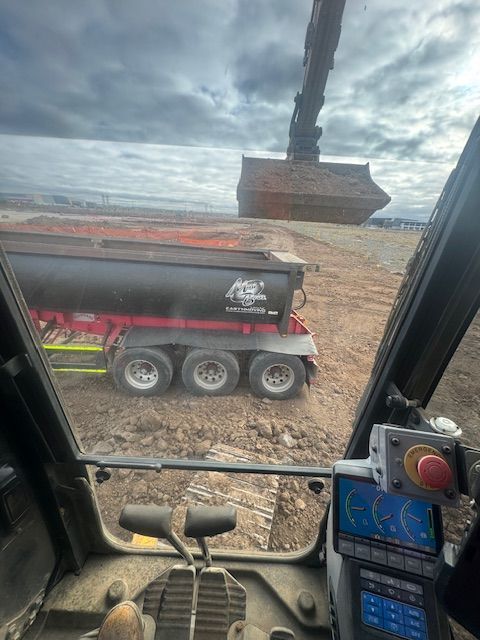 Cab view of an excavator loading gravel into a black dump trailer on a rocky construction site