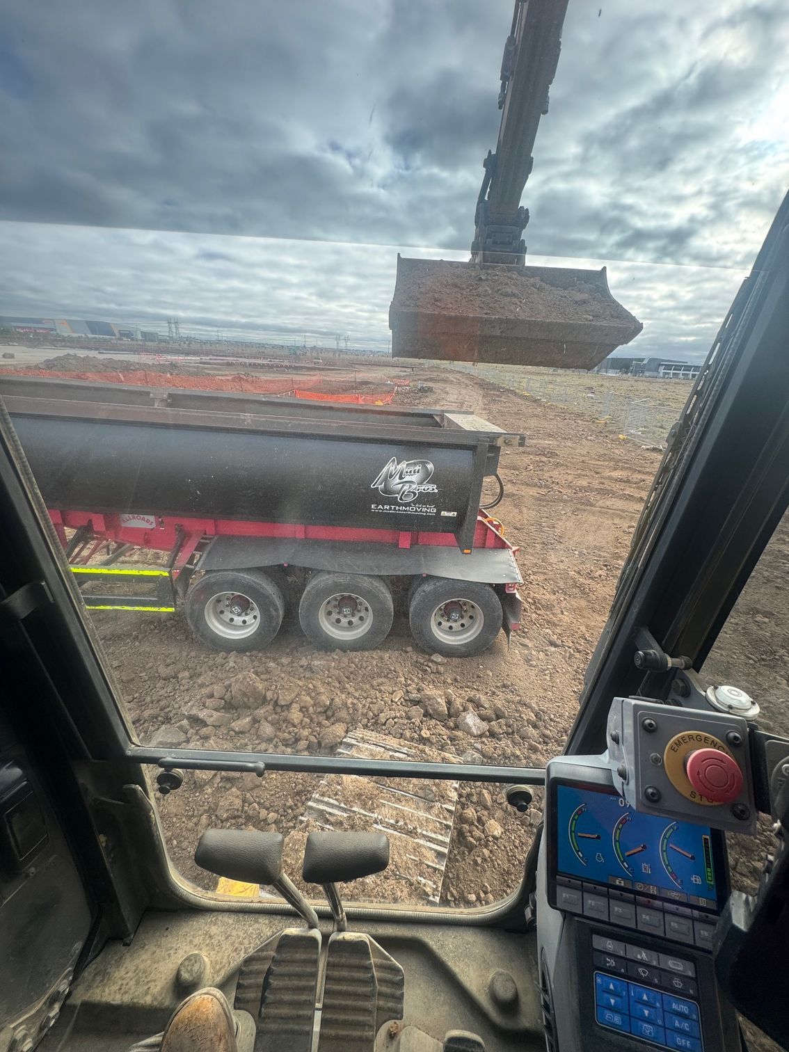 Inside a heavy machine cabin with an excavator loading a black dump truck on a muddy construction site under a cloudy sky.