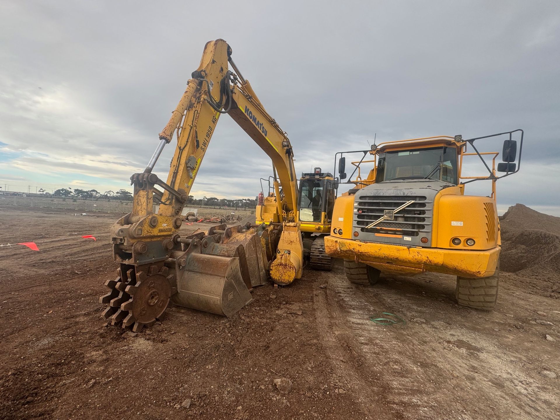 Yellow excavator and dump truck on a construction site, both facing forward, under a cloudy sky.