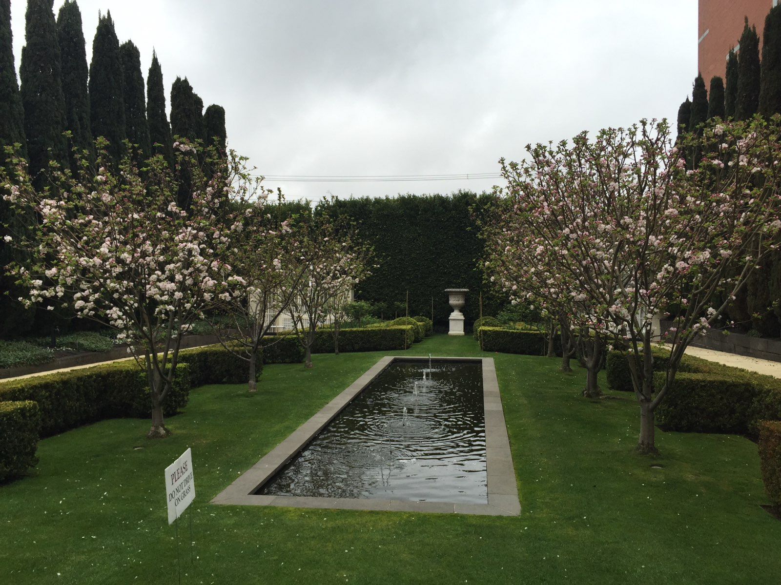 Formal garden with a rectangular reflecting pool, trimmed hedges, and blossoming trees under a cloudy sky