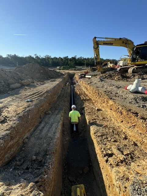 Worker in yellow vest in a deep trench at a construction site with an excavator nearby.