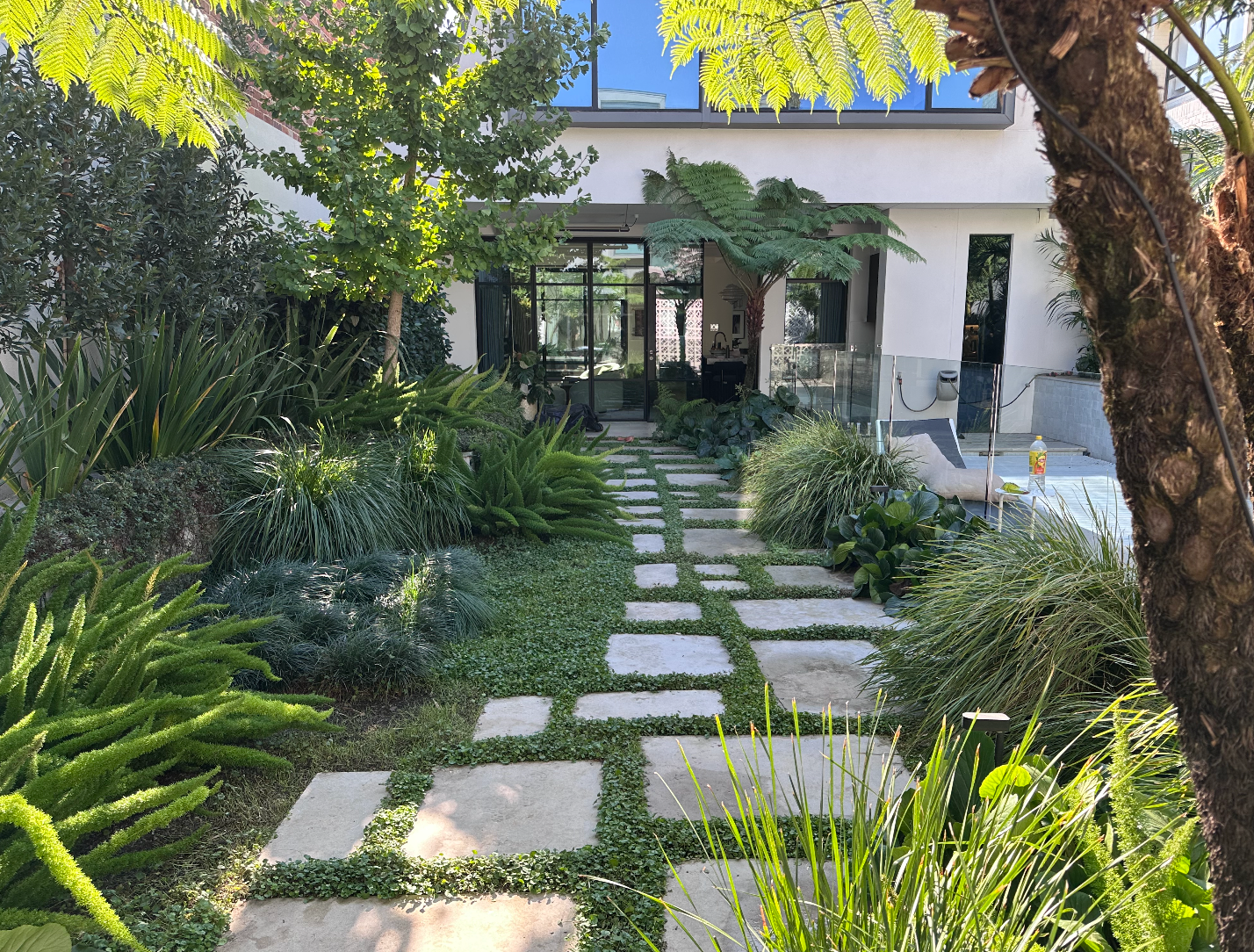 Stone path through lush garden to a white house with large windows and a patio.