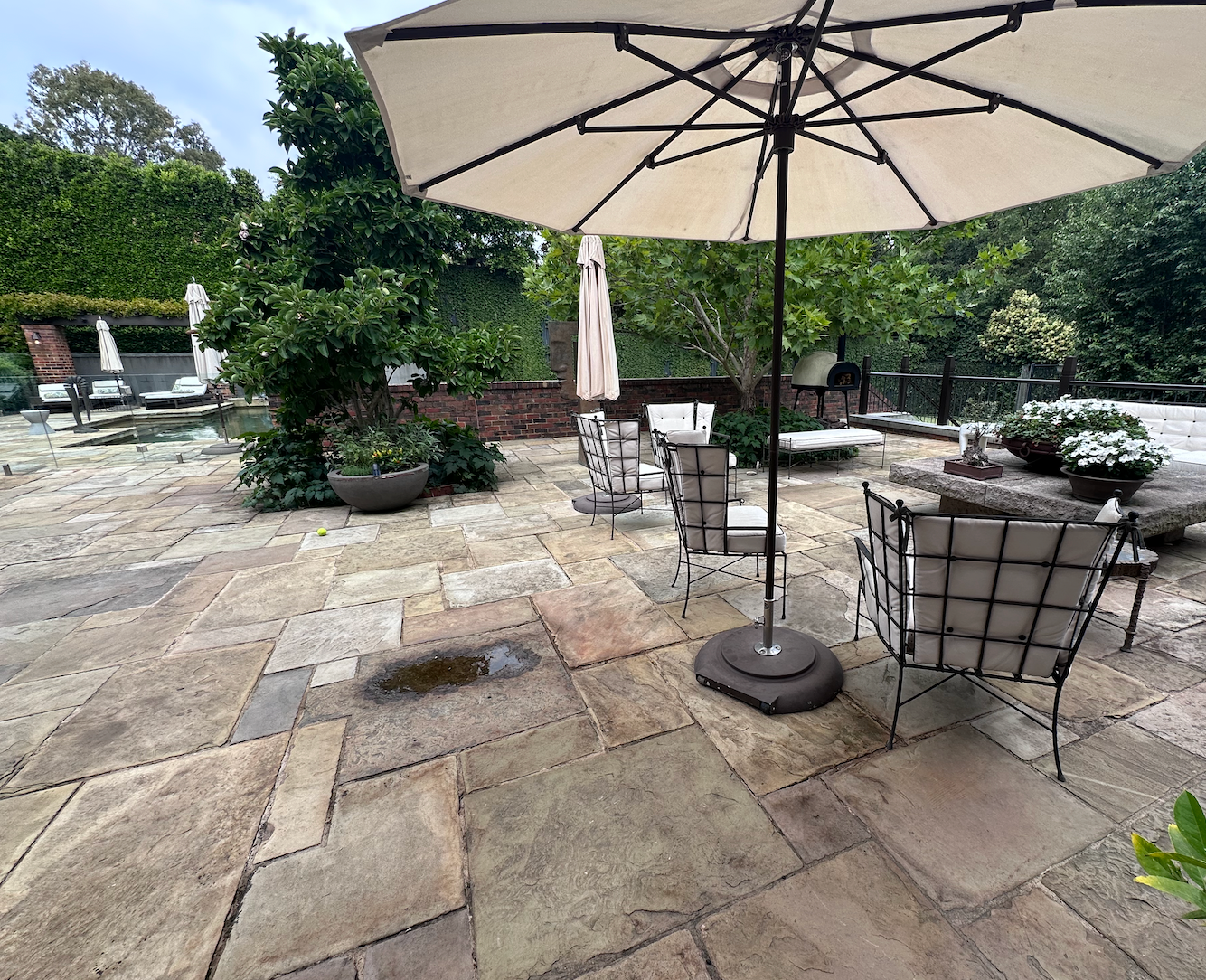 Patio with stone flooring, tables, chairs, and a large beige umbrella surrounded by greenery