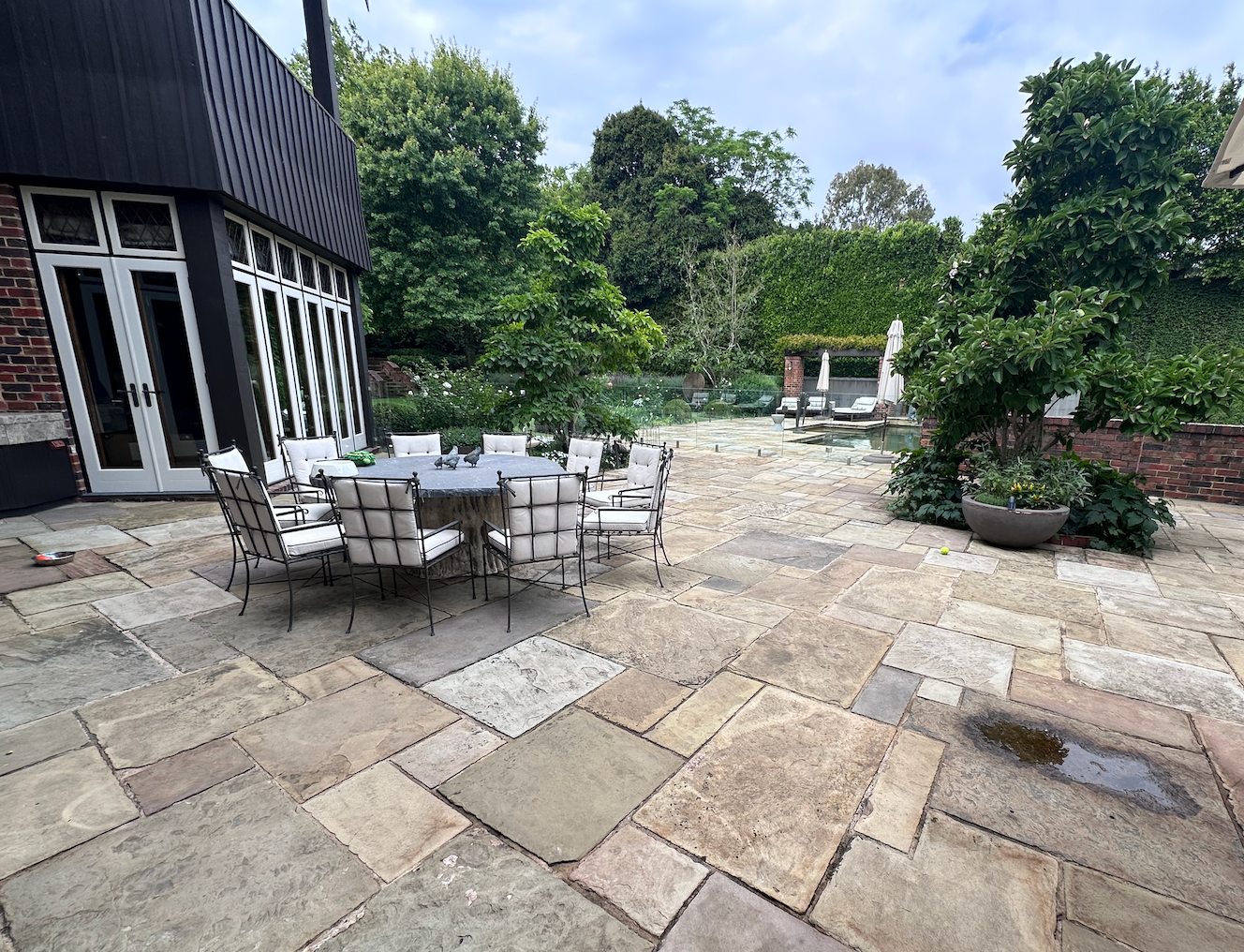 Patio with stone pavers, outdoor dining table and chairs beside a modern house, surrounded by greenery