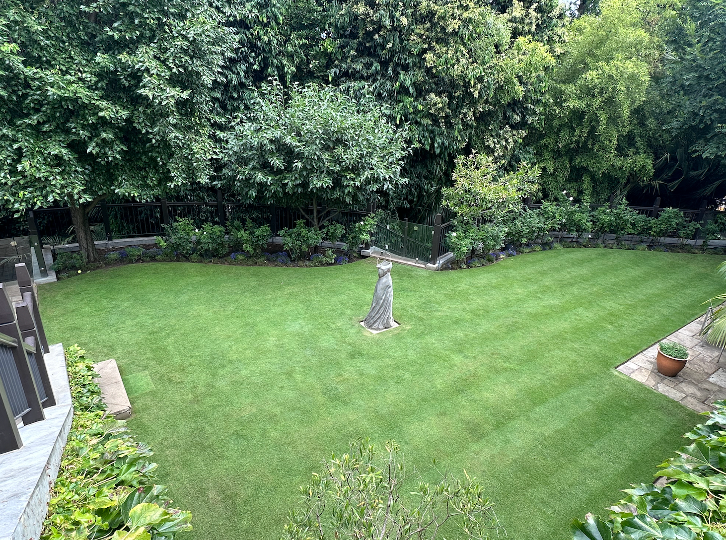 Lush green backyard lawn bordered by dense trees and shrubs, with a small stone statue near the center.