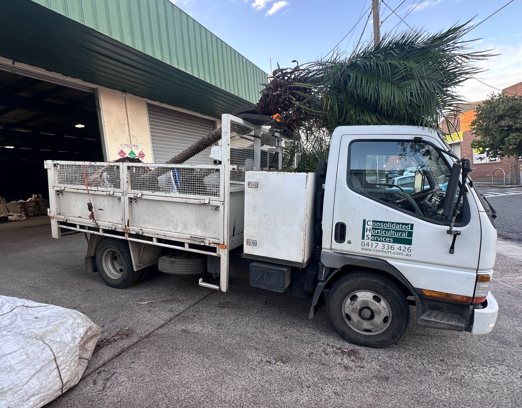 White utility truck parked beside a building with an open bay, loaded with green plant debris.