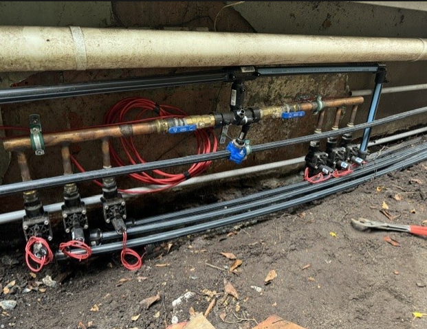 Pipe and valve assembly with black and red fittings mounted along a wall in an outdoor work area.