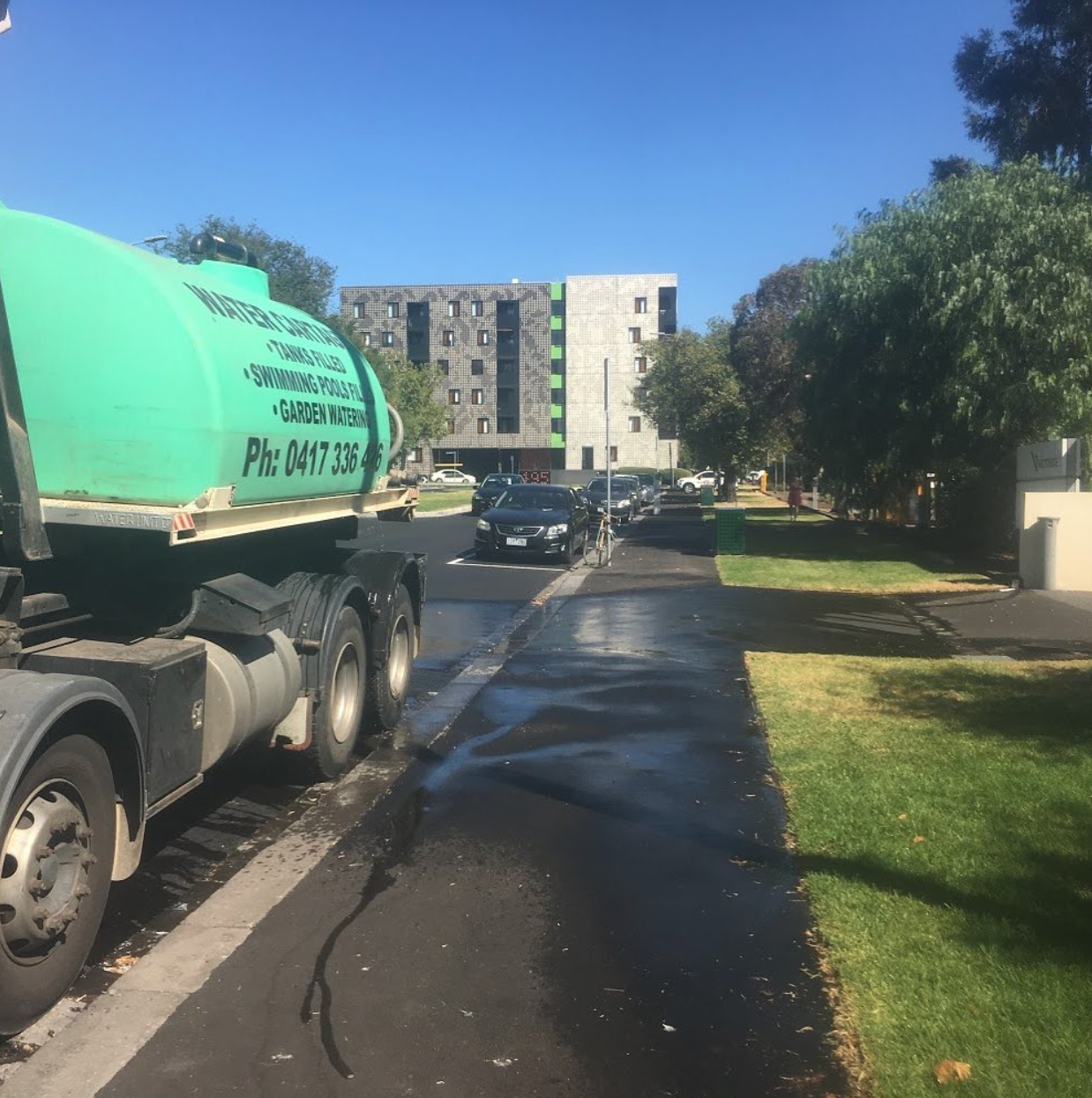 Green tanker truck on a wet driveway near apartment buildings and trees under a clear blue sky