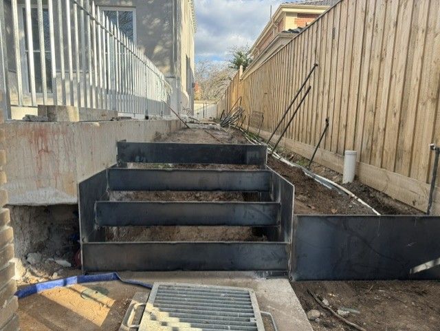 Backyard stairway under construction with black retaining walls beside a wooden fence and house wall