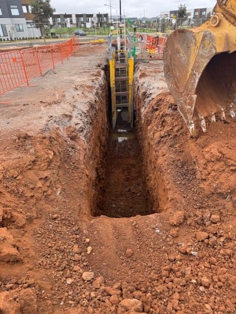 Construction trench with an excavator beside a deep, narrow excavation lined with orange safety fencing.