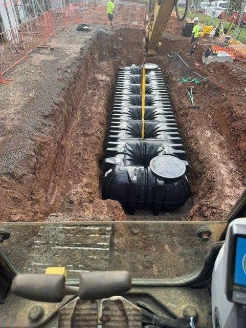 Construction site with a large black underground tank being lowered into a trench by excavator machinery.