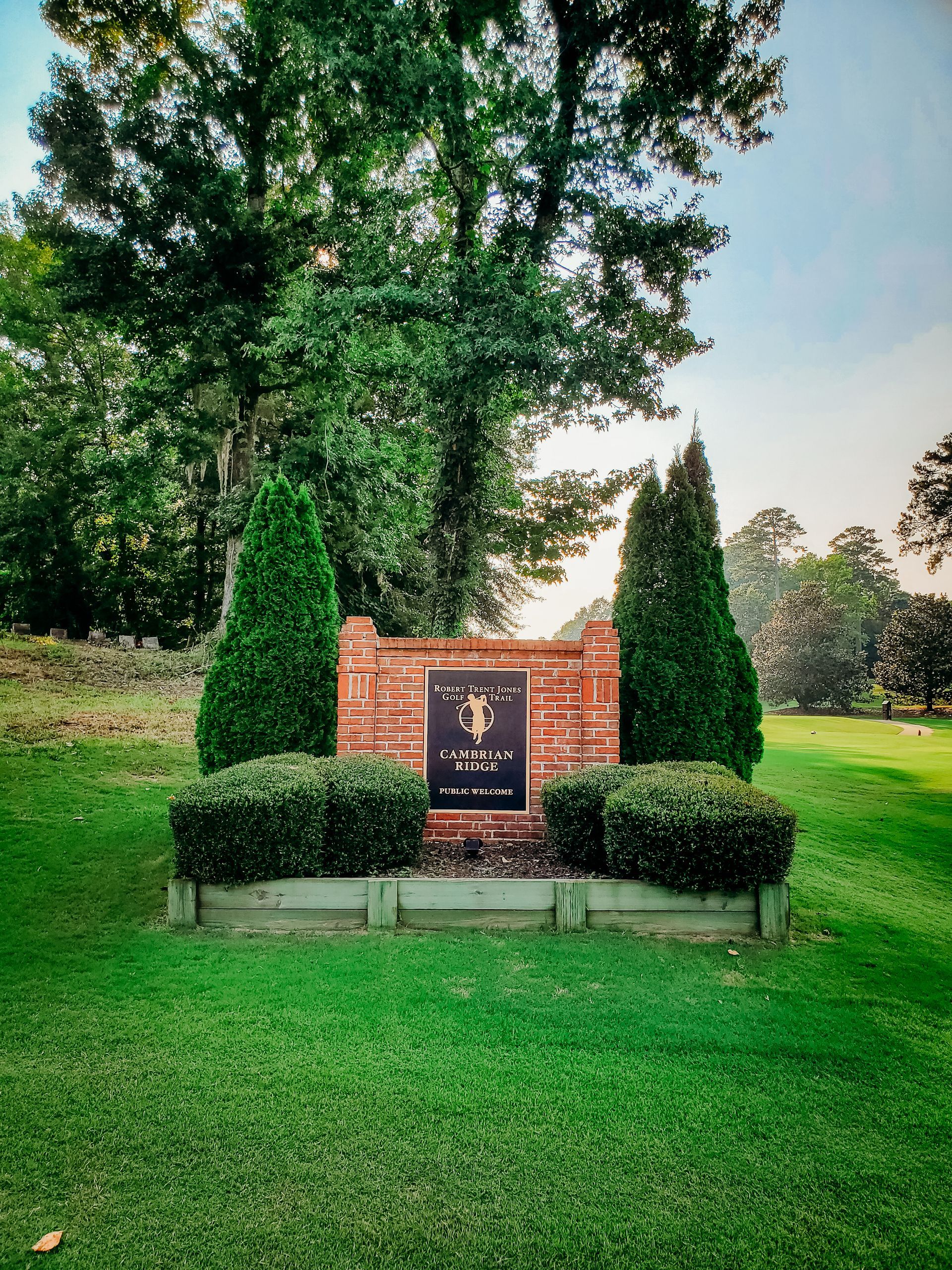Sign at a golf course entrance, brick and black plaque, flanked by two green trees.