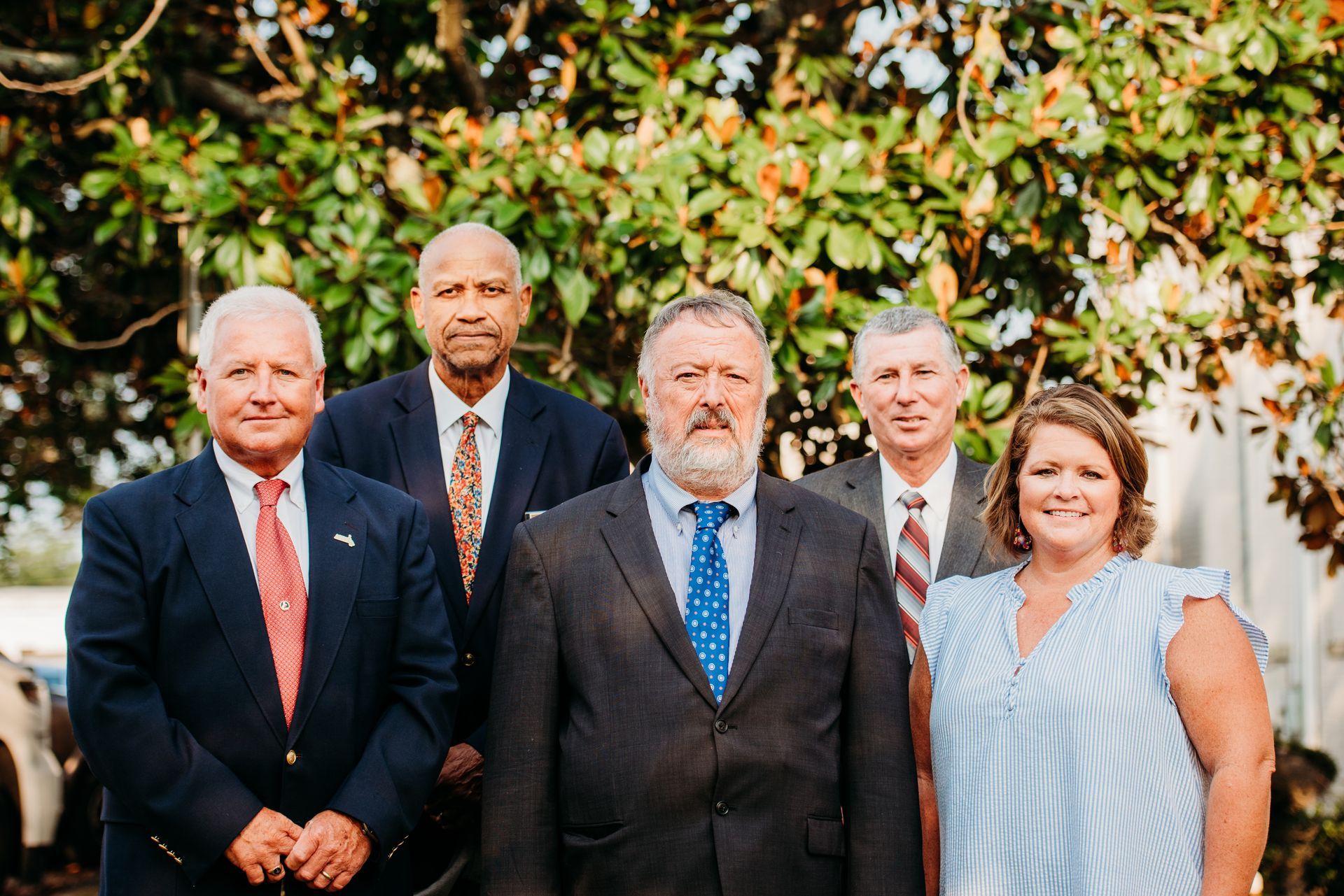 Group of six people in suits posing outside near green foliage.
