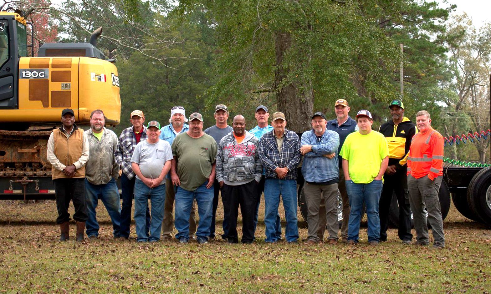Group of men standing in front of yellow construction equipment and trees.