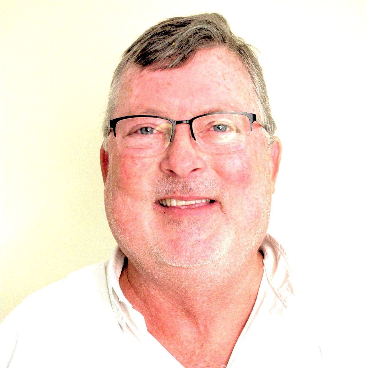 Man with glasses smiling, wearing a white shirt, against a pale background.