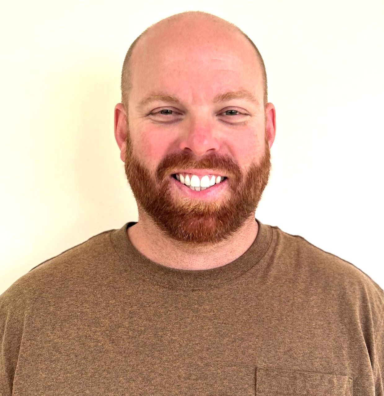 Bald man with a red beard smiles, wearing a brown shirt, against a pale yellow background.