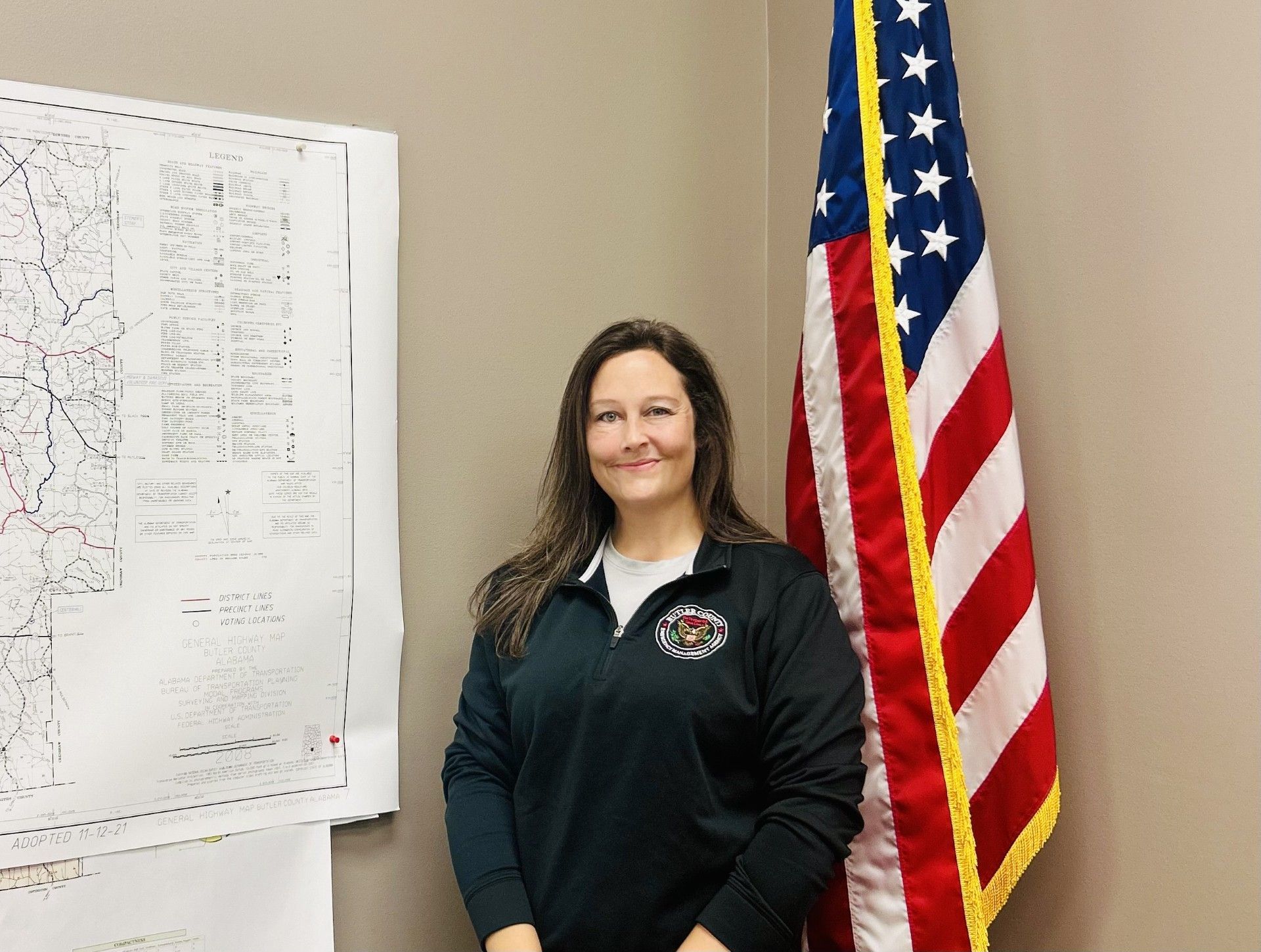 Woman standing beside an American flag and a map. She wears a black shirt, smiles in an office.