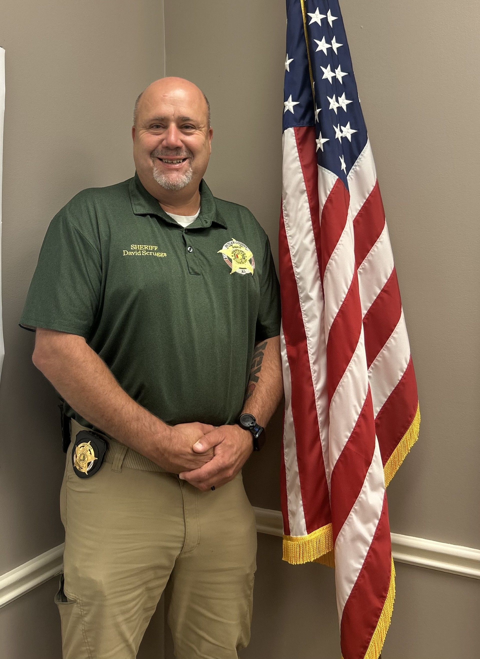 Man in green shirt, tan pants, badge, and watch, standing by American flag in room, smiling.