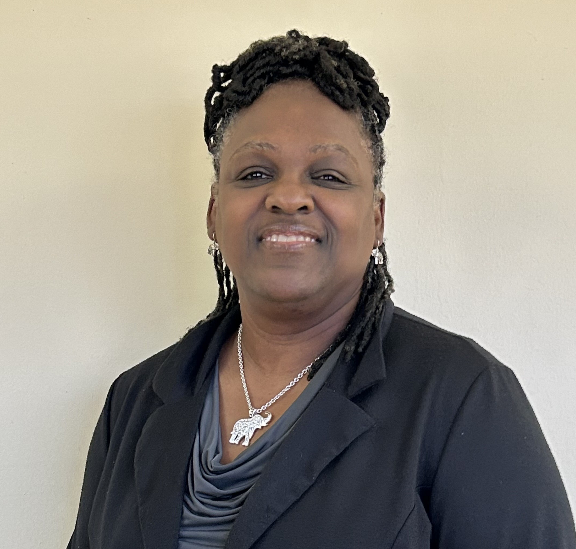 Woman in a blazer smiles, wearing a necklace. Natural lighting against a beige wall.