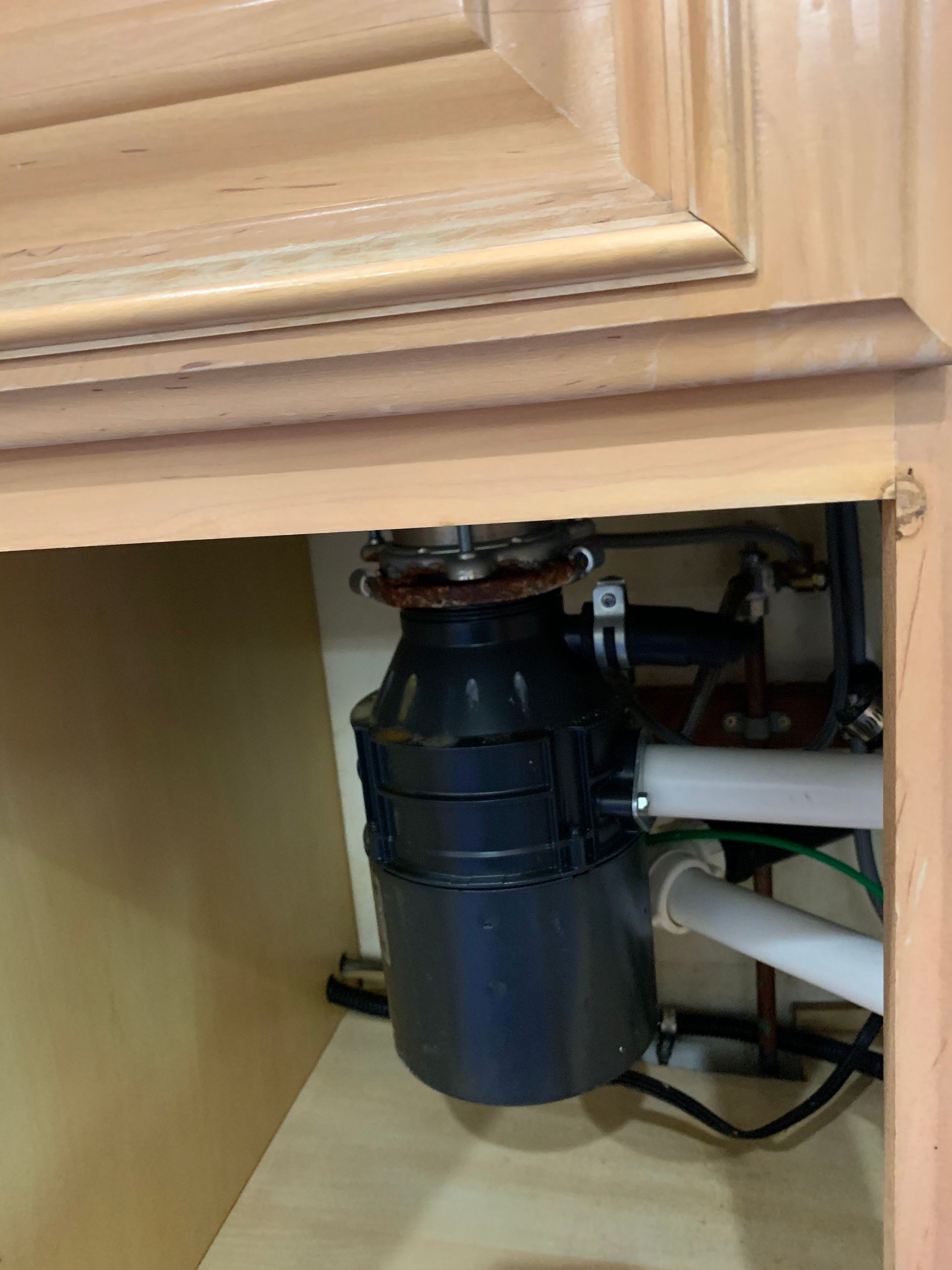 Under-sink garbage disposal unit with connected white plumbing pipes inside a light wood kitchen cabinet.
