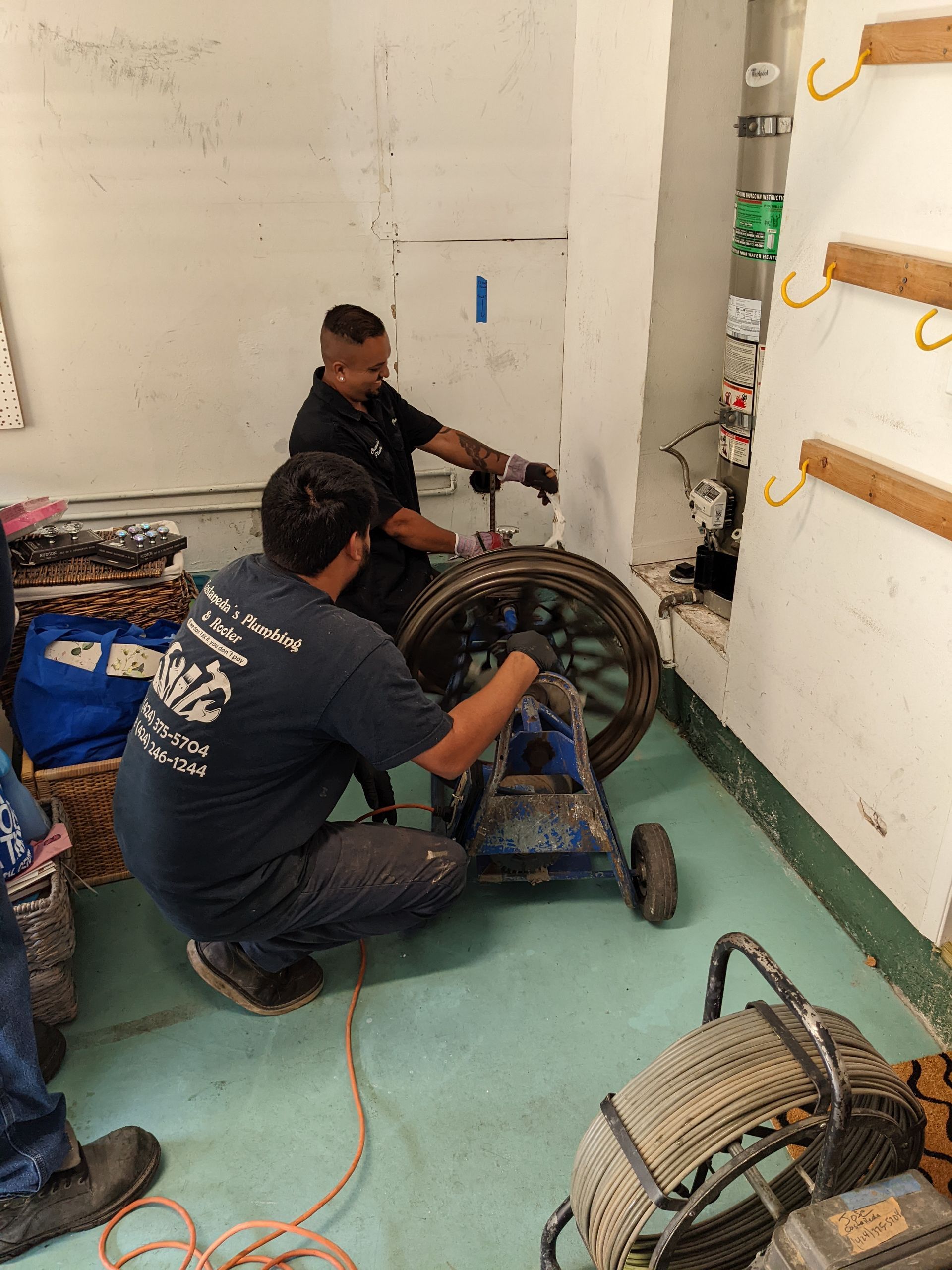 Two workers in a utility room use a drain-cleaning machine to service a pipe embedded in a wall.