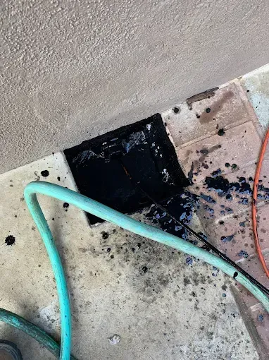 A garden hose is spraying water into an open, dark drain hole against a light-colored stucco wall and brick patio.