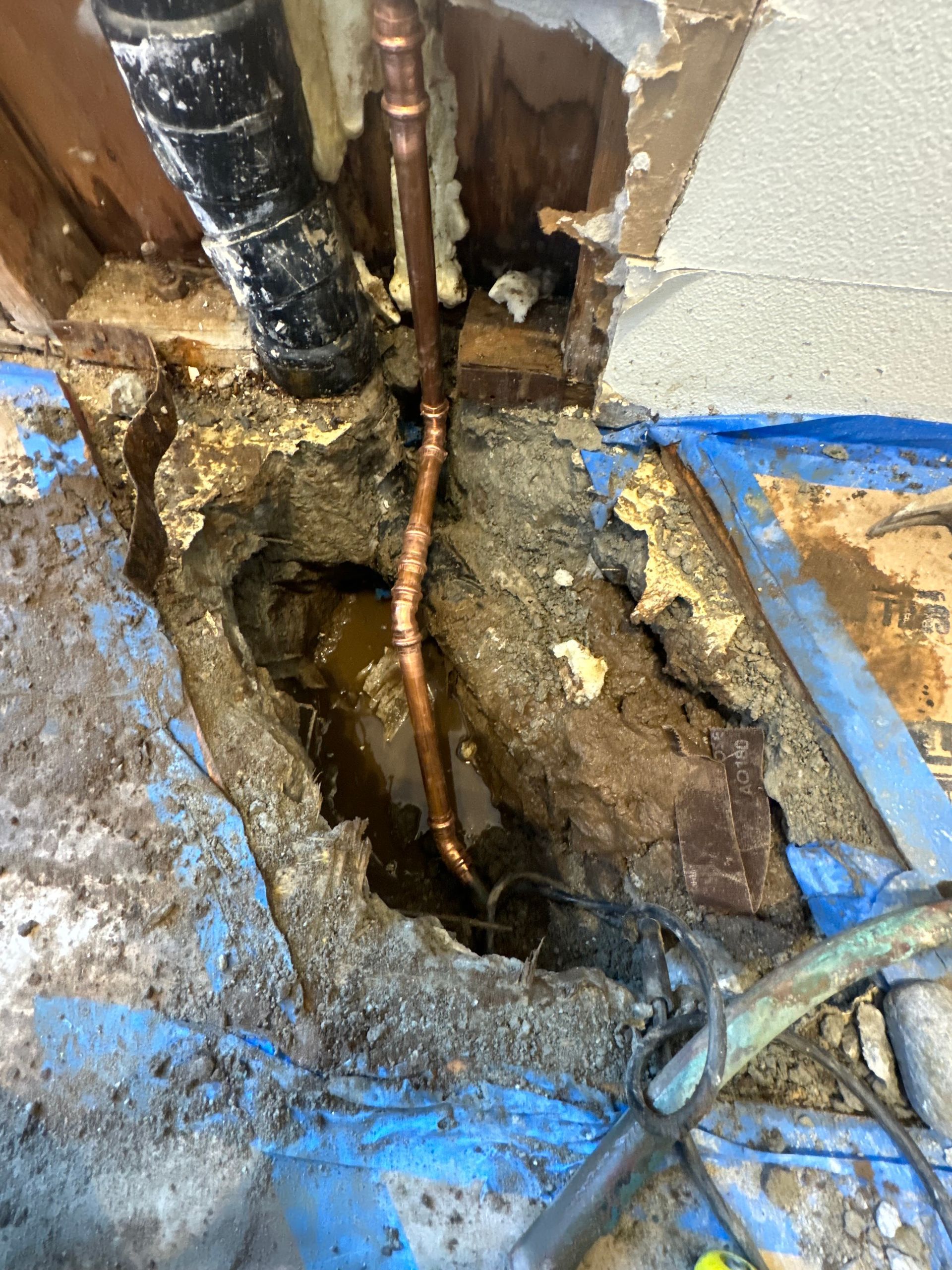 A hole broken through concrete flooring exposing soil, a black drain pipe, and new copper plumbing piping.