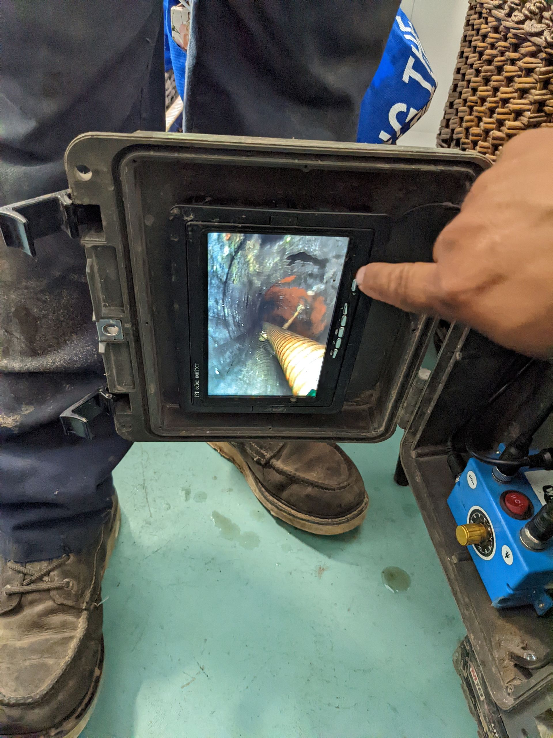 A person uses a handheld sewer inspection camera to view a dark, narrow pipe on a small digital monitor.