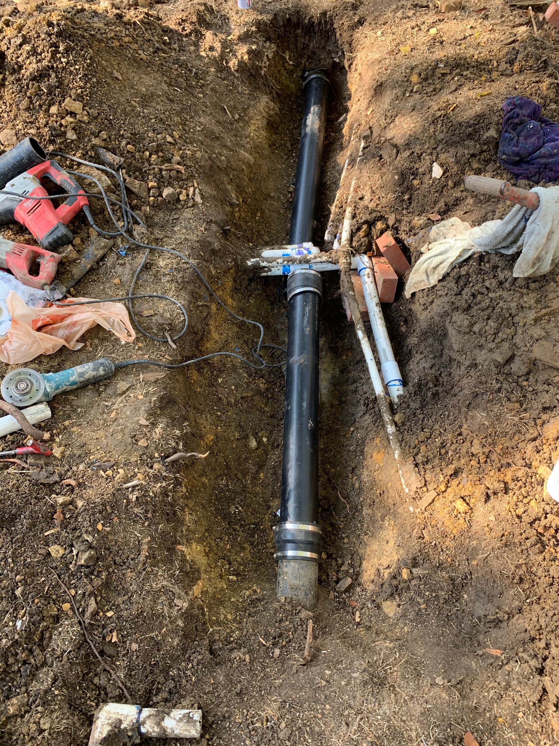 A black pipe installed in a narrow, dug-out trench in dirt, surrounded by scattered power tools and construction debris.