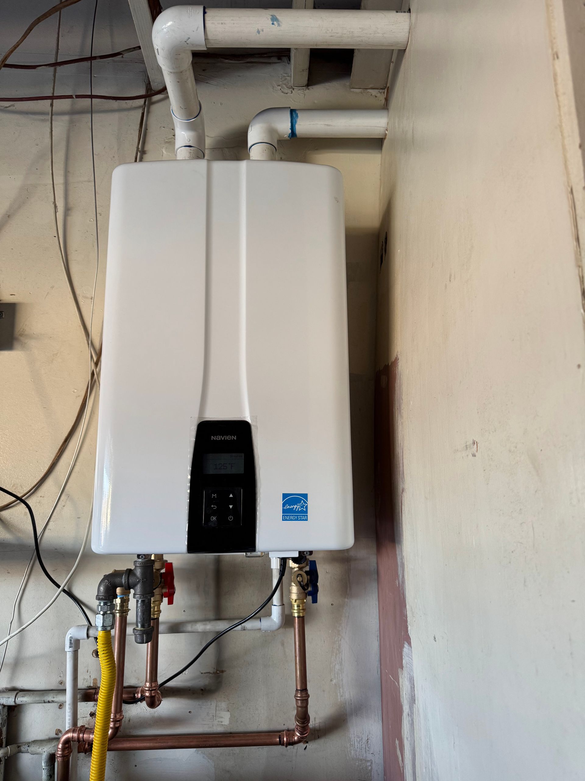 A white wall-mounted tankless water heater with connected pipes and wiring, installed against an unfinished interior wall.