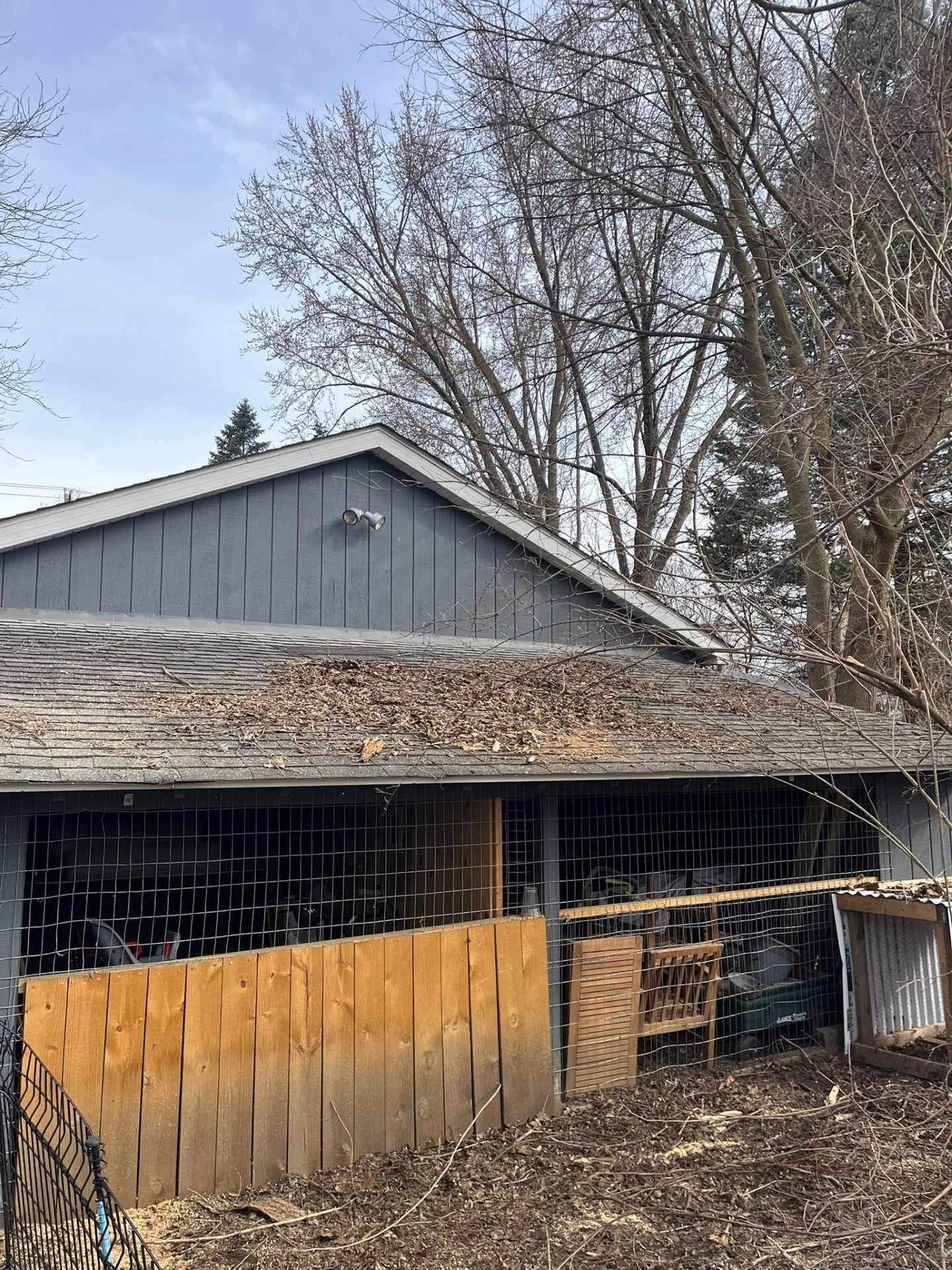 Weathered wooden shed with gray siding, leaf-covered roof, and wire fencing in a wooded yard