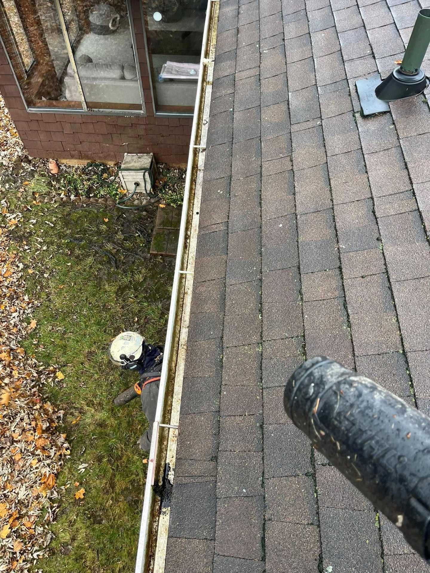 Roof edge beside a brick house and gutter, with a worker visible below in the yard