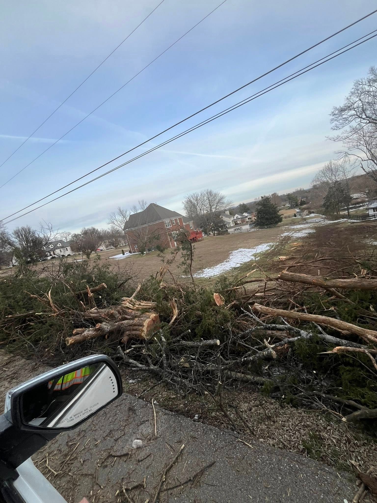 Storm-damaged roadside with fallen tree branches across a wet street, viewed from a car