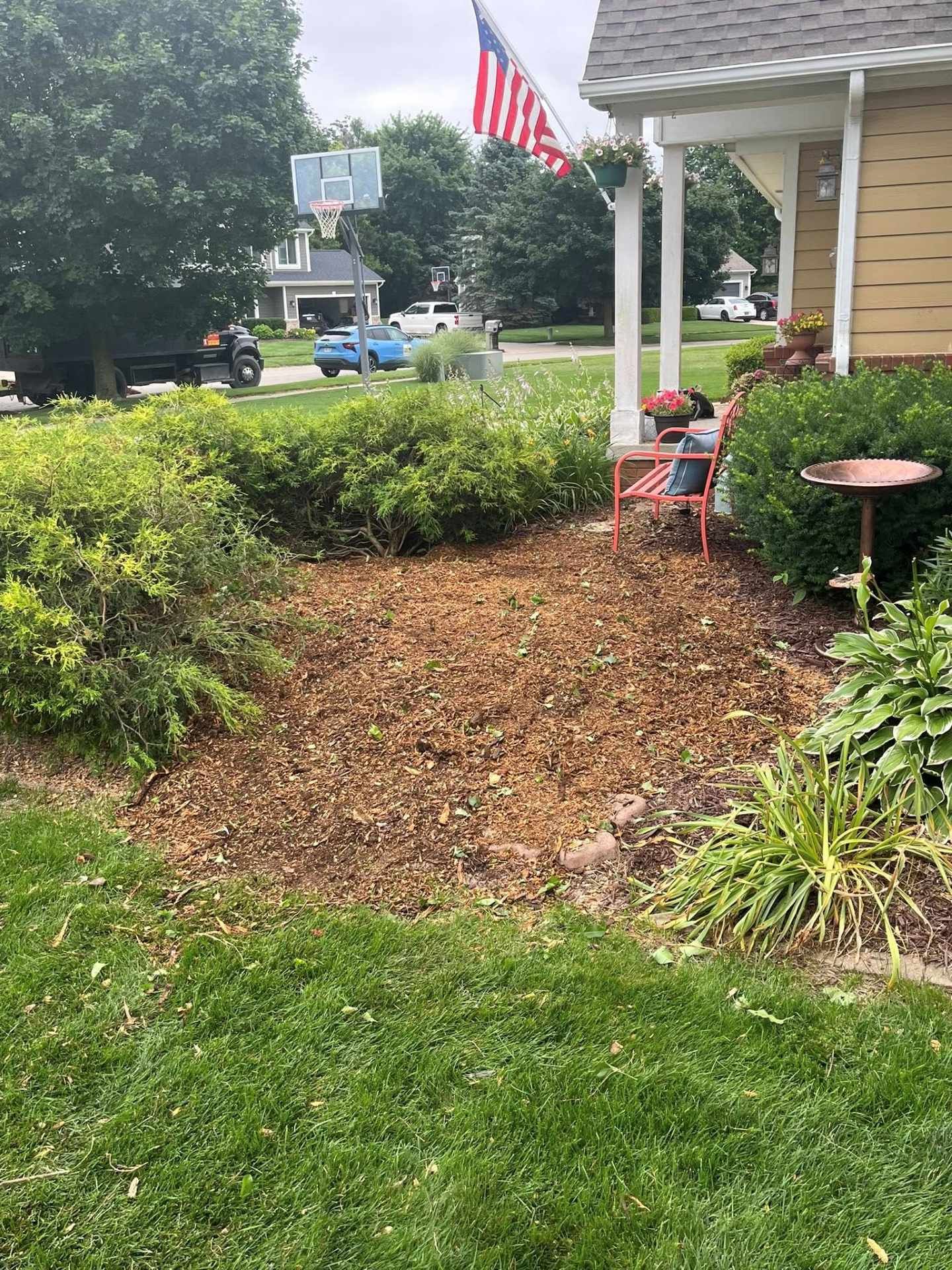 Front porch with red chair, mulch bed, shrubs, lawn, and an American flag by the house