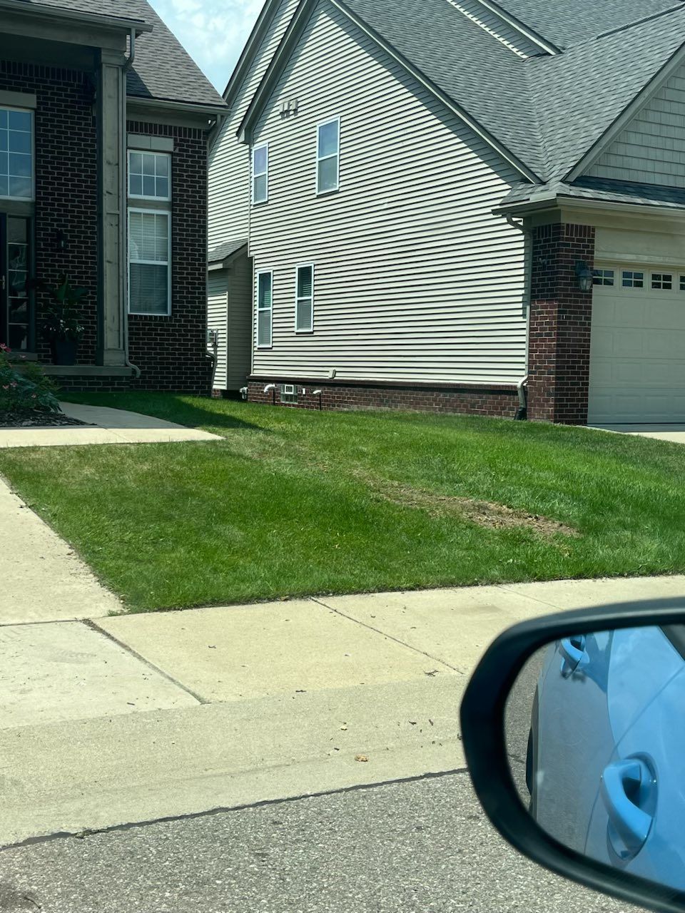 Suburban house exterior with light siding, brick accents, driveway, and front lawn viewed from a car.