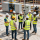 Obreros de la construcción con cascos y chalecos de seguridad forman un círculo en una obra con equipos cerca.