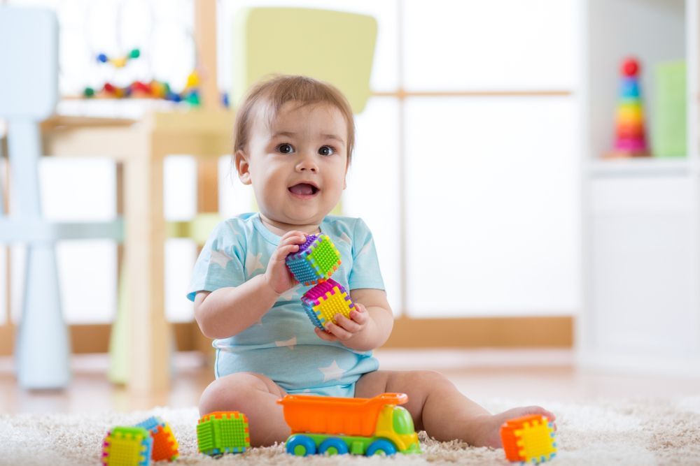 Baby playing with colorful toys on a rug, smiling in a bright room.