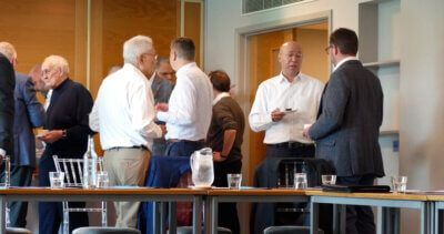 A group of men are standing around tables in a room talking to each other.